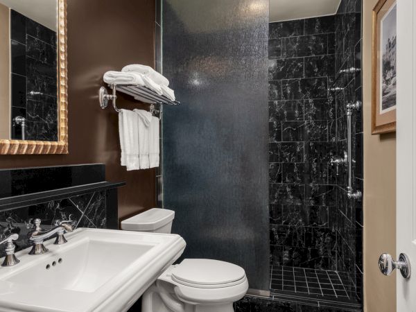 A modern bathroom with a white sink, toilet, and a black-tiled shower. There's a towel rack with folded towels above the toilet.
