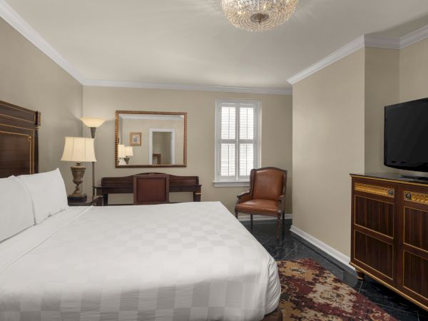 A cozy bedroom with a white bed, wooden furniture, TV, leather chair, and elegant chandelier, creating a warm, inviting atmosphere.