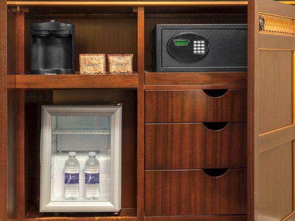 A wooden cabinet has a coffee maker, two coffee packs, a safe, a mini refrigerator with two water bottles.