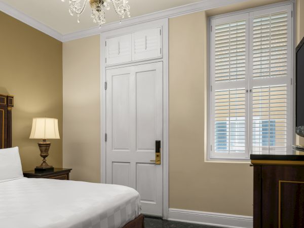 A hotel room with a bed, wooden headboard, nightstand, lamp, TV, door, window with shutters, and chandelier on the ceiling.