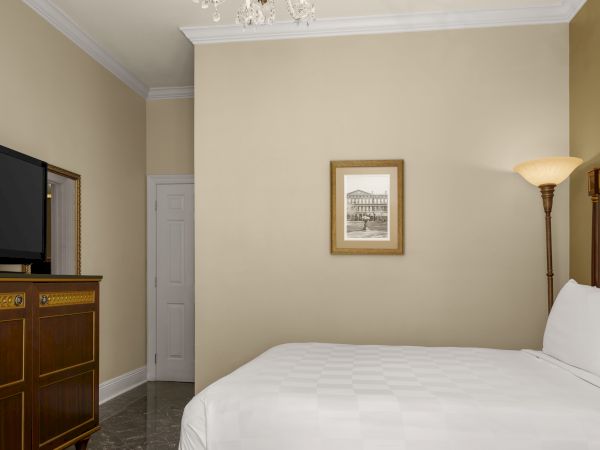 A hotel room with a bed, framed artwork, TV, and a floor lamp, decorated in neutral tones, offering a cozy atmosphere.
