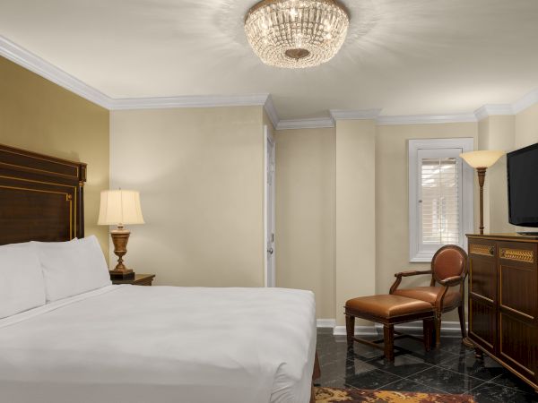 A hotel room with a large bed, ornate chandelier, wooden furniture, and a TV on a cabinet. There's a soft lamp near the bed.
