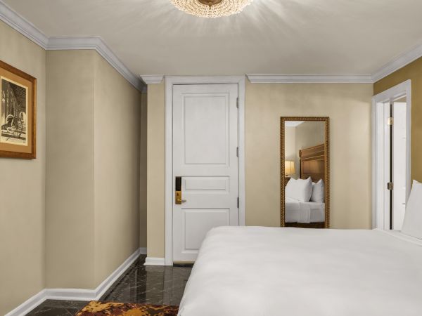 A hotel room features a bed, mirror, artwork, and a white door with a modern light fixture on the ceiling.