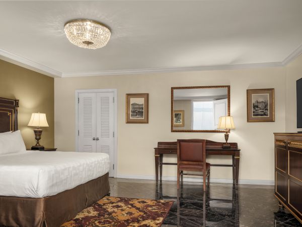 A neatly arranged hotel room with a bed, ornate desk and chair, large mirror, paintings, TV, and elegant lighting on the ceiling.