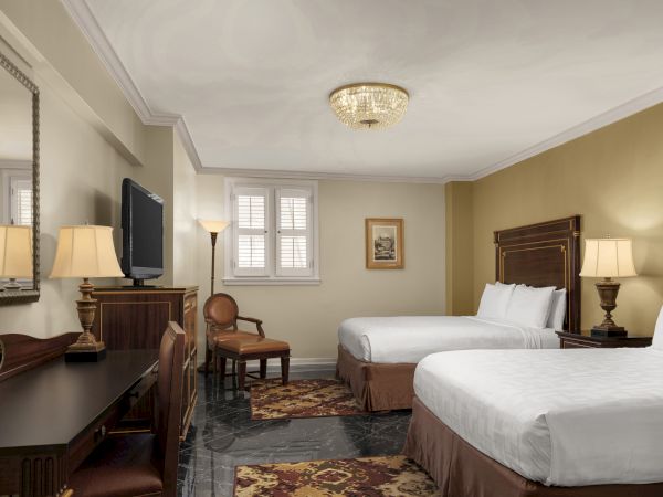 A hotel room with two beds, a desk, a chair, a TV, lamps, a window, and framed wall art, decorated in a classic style.
