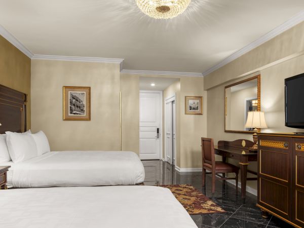 This image shows a hotel room with two beds, a desk, chair, TV, lamp, decorative mirror, and elegant lighting fixture on the ceiling.