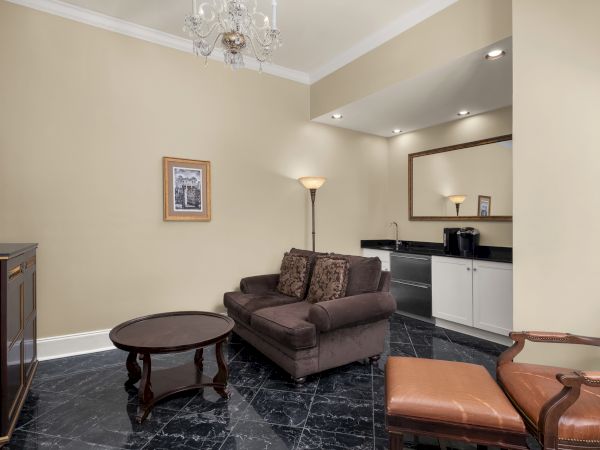 A living area with a brown sofa, armchair, and coffee table. There's a TV, floor lamp, chandelier, artwork, and a kitchenette with a mirror.