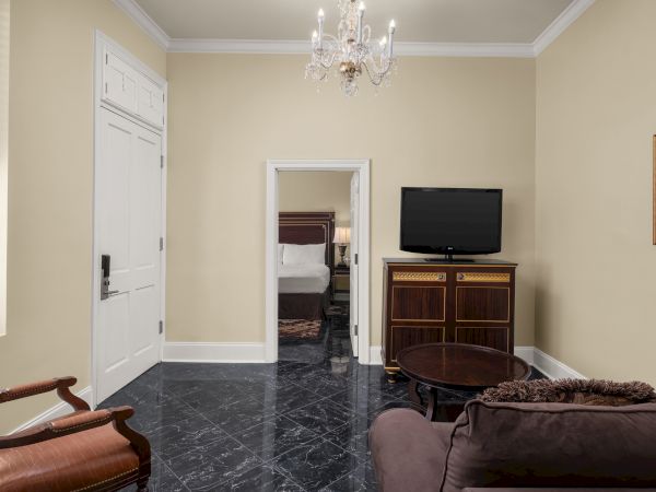 A living room with a chandelier, TV on a cabinet, sofa, chair, round table, and a view into a bedroom with a bed and lamp.