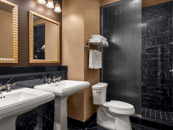 A luxurious bathroom with dual sinks, mirrors, toilet, black marble tiles, and a walk-in shower with towels neatly arranged above.
