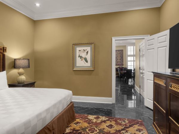 A bedroom with a large bed, nightstand with a lamp, framed art, and a dresser with a TV. An open door leads to another room.