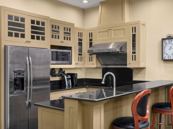 A modern kitchen with beige cabinets, stainless steel appliances, a black countertop, and bar stools. A wall clock hangs on the wall.