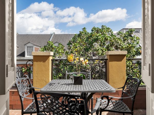 A patio scene with a metal table and chairs overlooking a garden and rooftops under a blue sky with clouds.