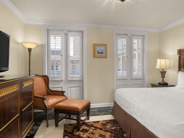 A cozy room with a bed, armchair, TV, and lamps. White shutters cover the windows. Artwork hangs on the beige walls.