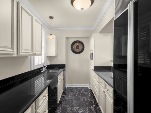 A modern kitchen with cream cabinets, black countertops, a wall clock, pendant light, and black marble floor.