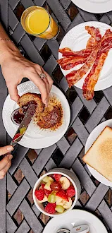A brunch table spread with bacon, eggs, toast, granola, fresh fruit cups, juice, and coffee on a tiled surface.